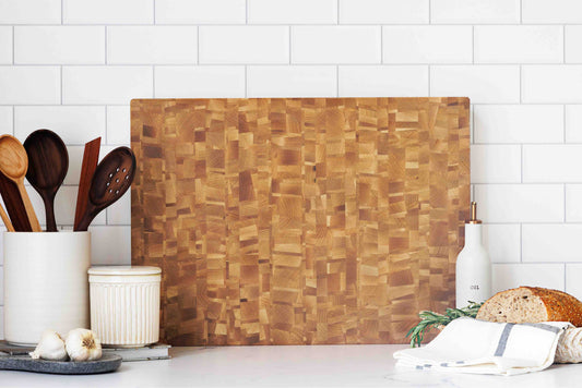 Natural maple cutting board styled in a modern kitchen setup with utensils, bread, and garlic nearby.